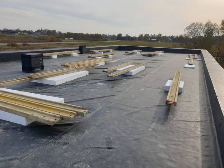 Płaski dach pokryty czarną membraną, z przygotowanymi legarami i styropianowymi podkładkami, widok z góry na tle jesiennego krajobrazu.