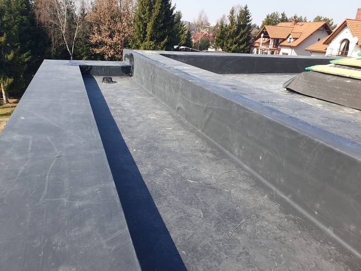 Płaski dach pokryty czarną membraną hydroizolacyjną z widocznymi elementami odprowadzającymi wodę i fragmentami zabudowy w tle.