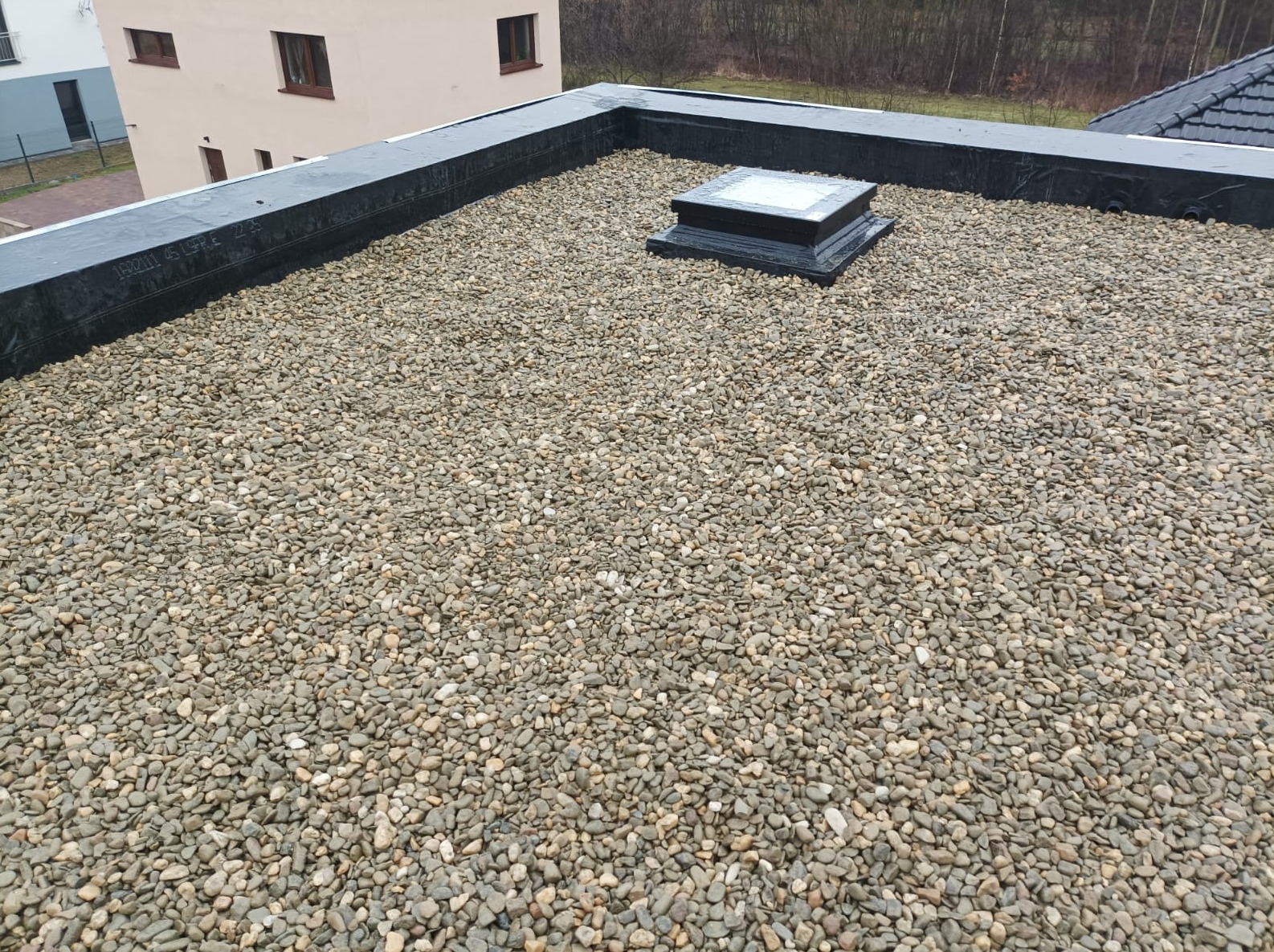 Płaski dach pokryty warstwą drobnych, okrągłych kamieni, z czarnym obramowaniem i świetlikiem w centralnej części, na tle zabudowań i drzew.