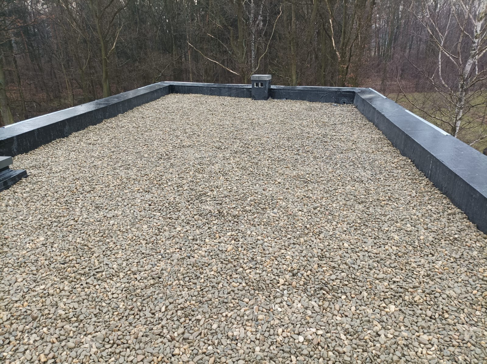 Płaski dach pokryty drobnym żwirem, otoczony czarną, gumową obwódką, z kominem wentylacyjnym w tle i widokiem na drzewa bez liści.