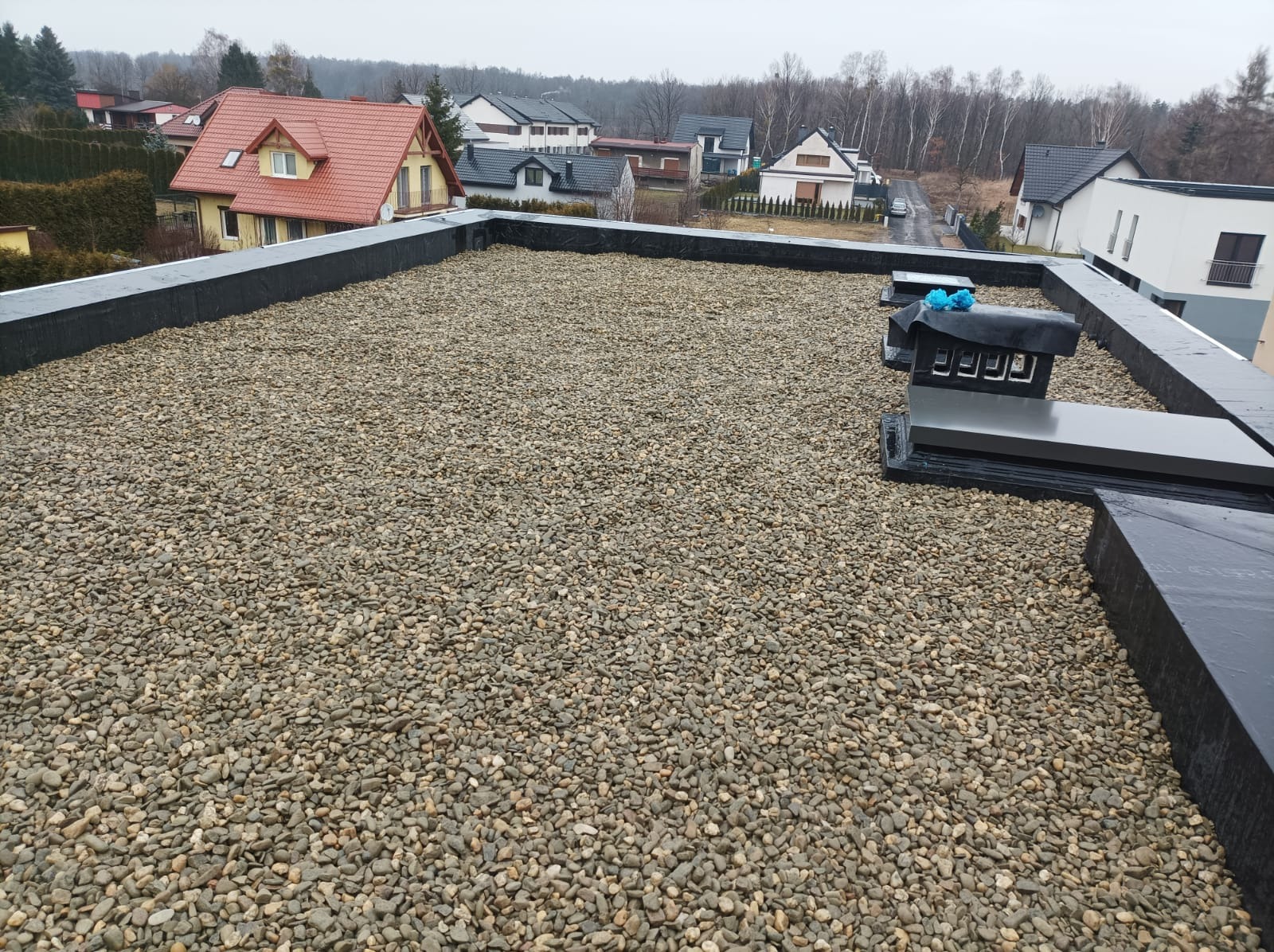 Płaski dach pokryty drobnymi kamieniami, z widocznymi kominami wentylacyjnymi i obróbką blacharską, na tle zabudowy jednorodzinnej w pochmurny dzień.