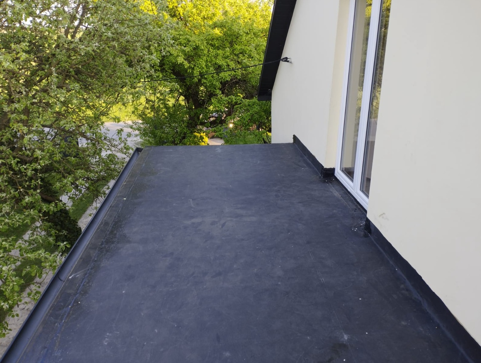 Widok z góry na płaski dach pokryty czarną membraną bitumiczną, z widoczną ścianą budynku w kremowym kolorze i oknem PCV, oraz bujną zielenią drzew w tle.