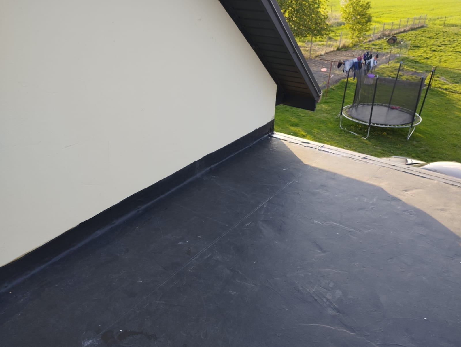 Czarny, płaski dach pokryty papą na tle domu z jasną elewacją, w oddali zielony trawnik z trampoliną i suszącym się praniem.