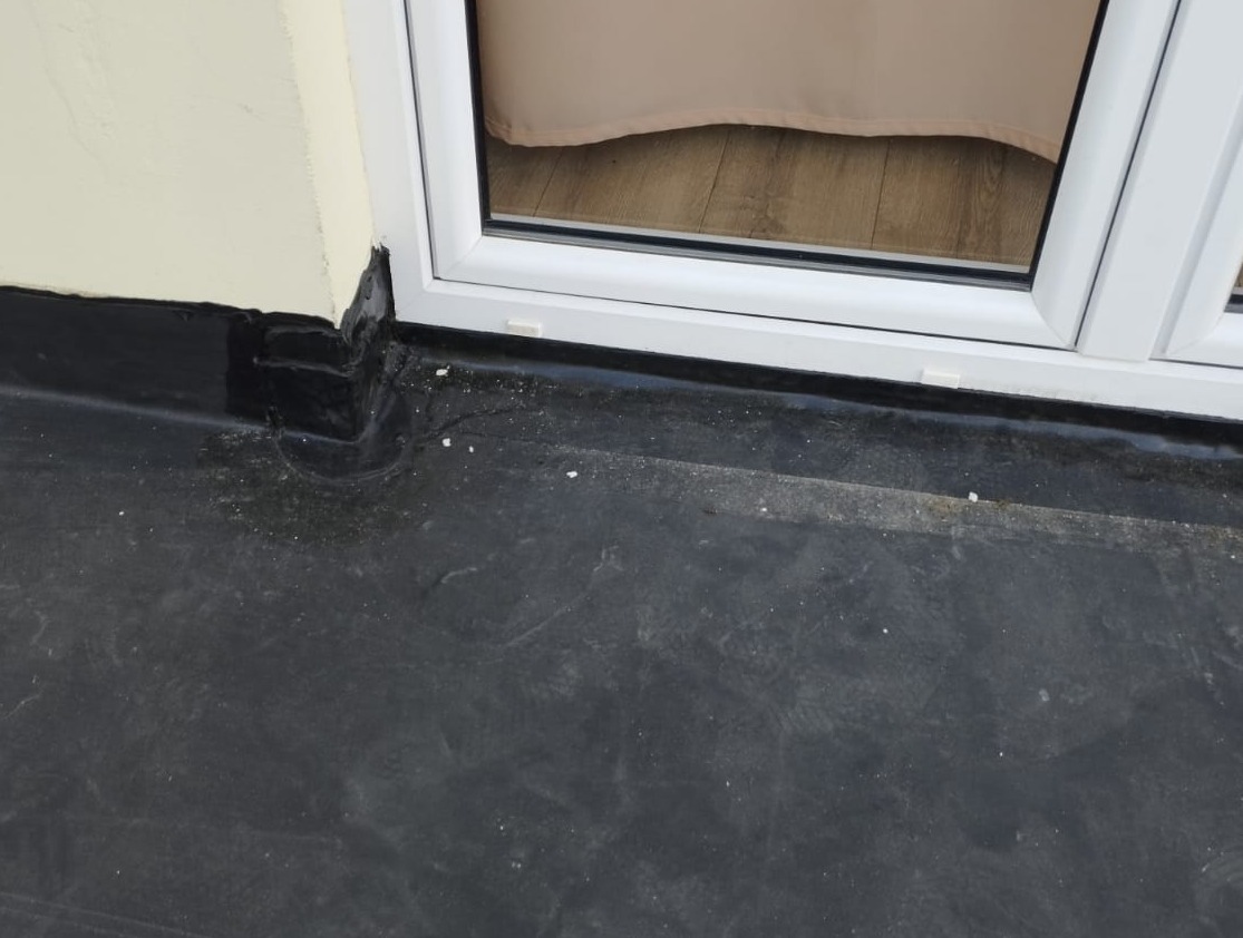 Płaski dach pokryty czarną membraną bitumiczną, z widocznym narożnikiem przy ścianie i oknem balkonowym z białymi ramami.