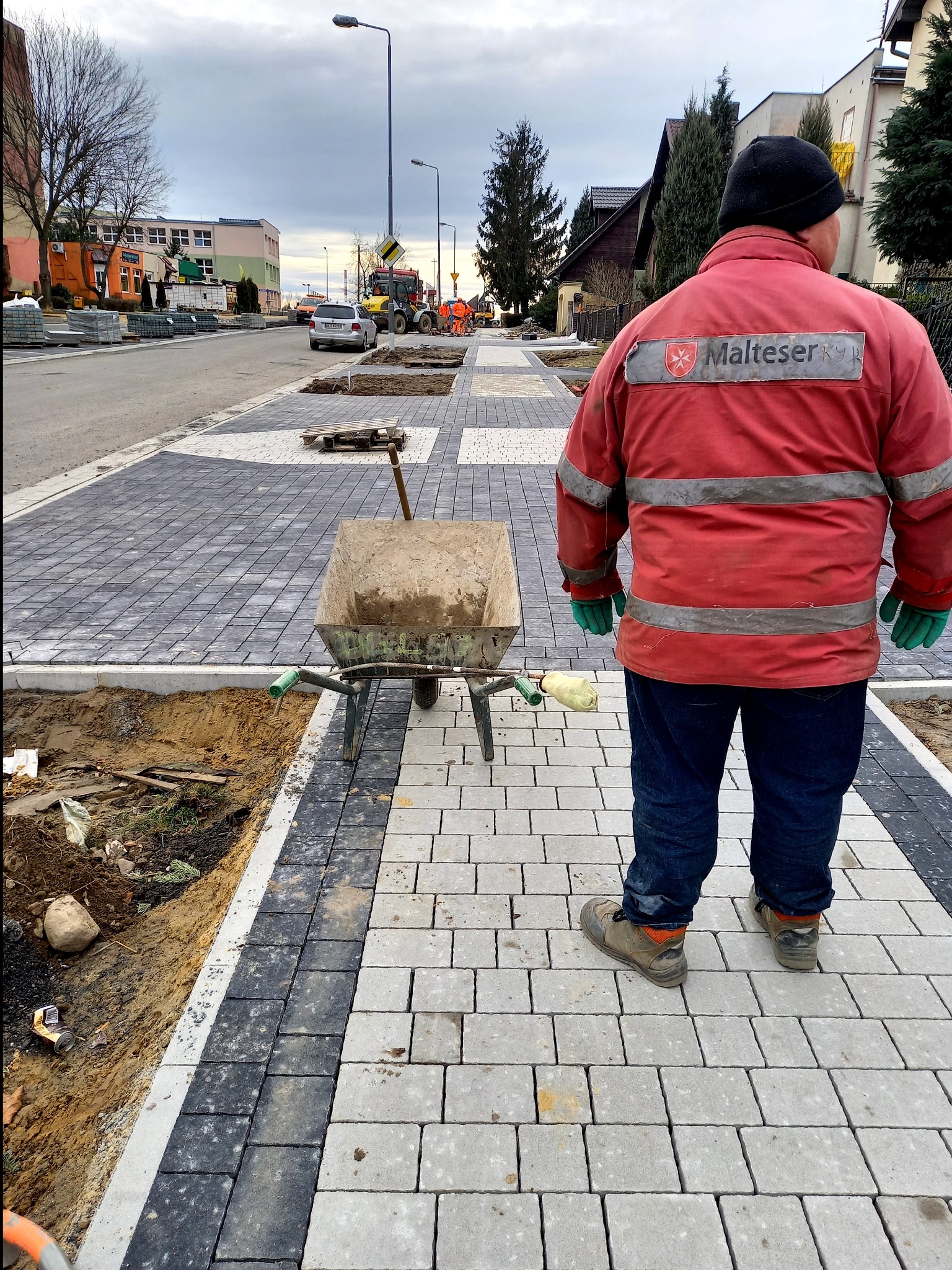 Układanie szarej kostki brukowej na chodniku z widocznym pracownikiem w czerwonej kurtce z logiem Malteser, taczką z zaprawą i fragmentem wykopu pod obrzeża.