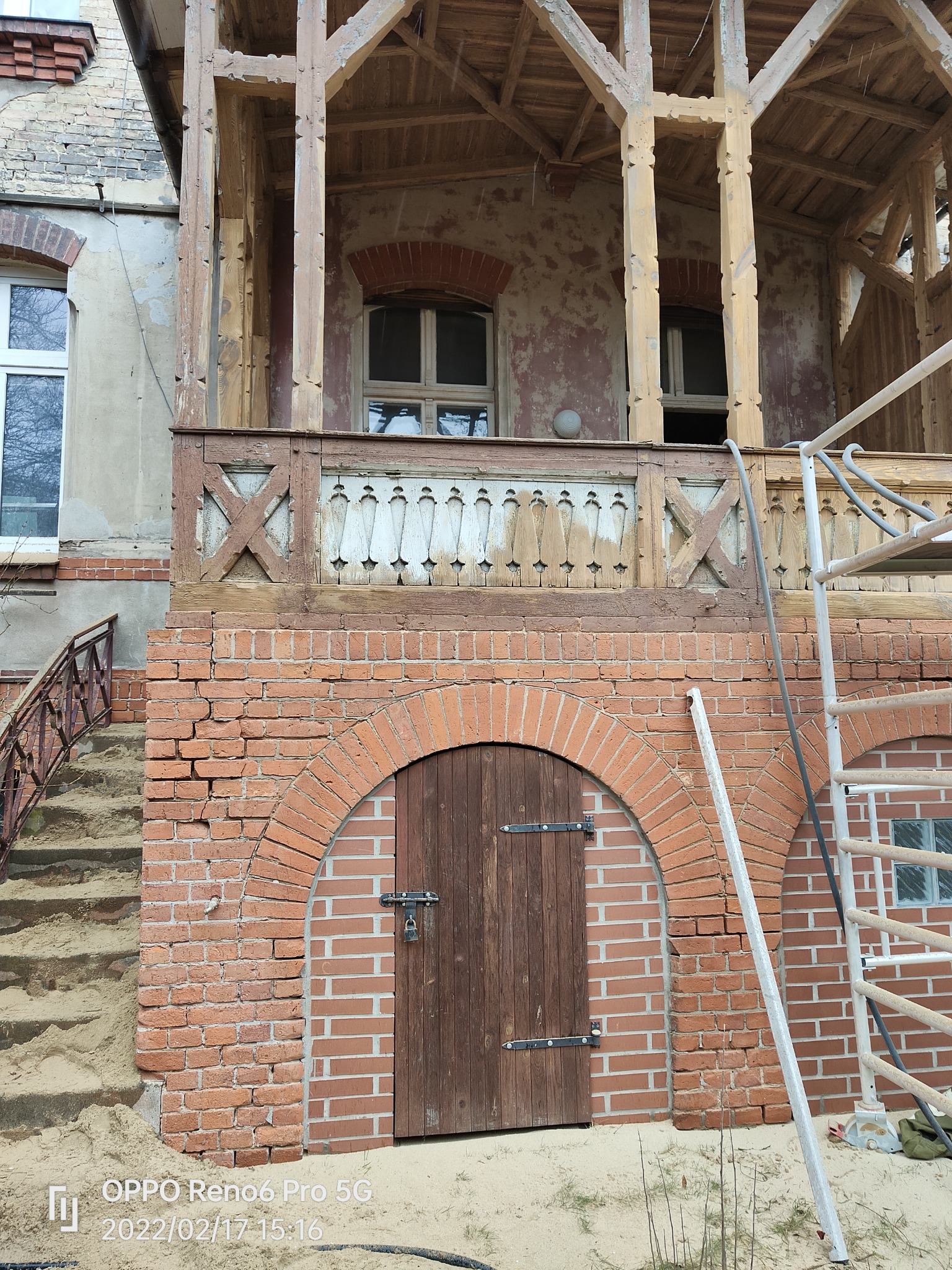 Drewniana weranda z ceglanym łukiem nad drzwiami, widoczne ślady przygotowania do renowacji, częściowo usunięta stara farba, rusztowanie obok.
