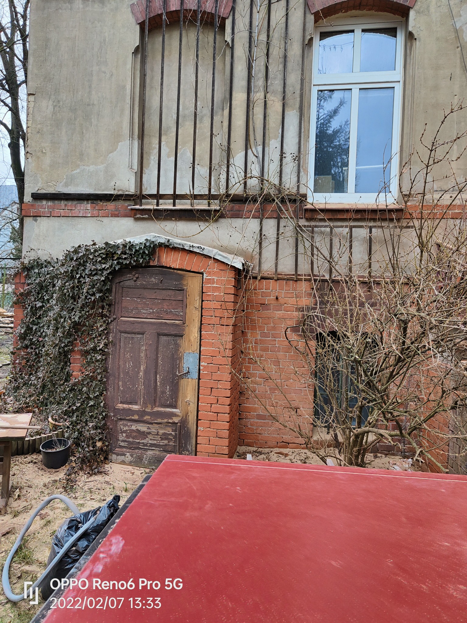 Stara, drewniana brama obrośnięta bluszczem, z widocznymi śladami odpryskującej farby, przylegająca do ceglanego budynku. W tle okno z białymi ramami i metalowe kraty. Na pierwszym planie czerwona,...