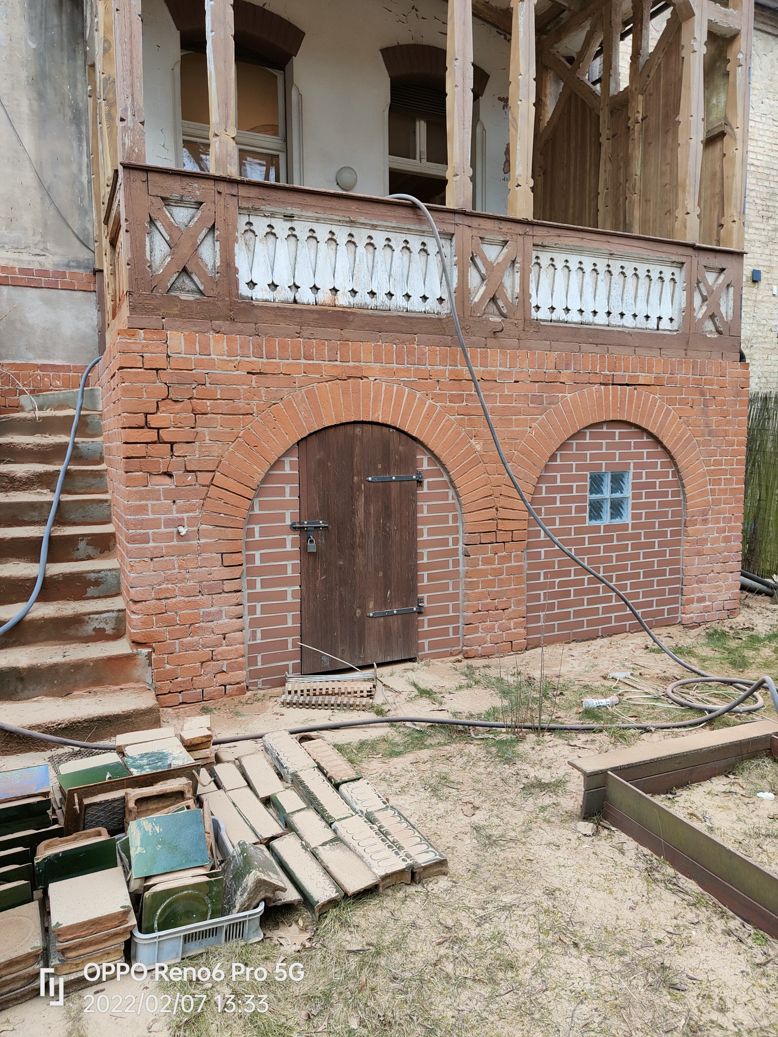 Piaskowana i pomalowana na biało balustrada balkonu z drewnianymi zdobieniami, ceglana podmurówka z łukowymi wejściami i schodami, przygotowane do ułożenia płytki na ziemi.