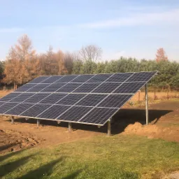 Panel fotowoltaiczny na metalowej konstrukcji w ogrodzie, świeżo po montażu, z widoczną ziemią i trawą, w tle drzewa z jesiennymi liśćmi.