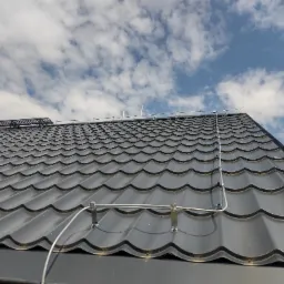 Szara blachodachówka z zamontowaną instalacją odgromową i elementami systemu solarnego w tle, na tle błękitnego nieba z chmurami.