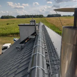 Widok z góry na zamontowane na dachu kominki wentylacyjne z czarnymi rurami i metalowymi obejmami, obok drabinka kominiarska, w tle pole uprawne i błękitne niebo z chmurami.