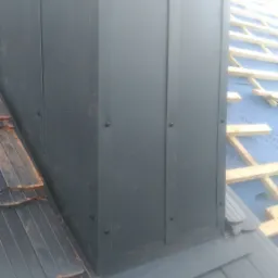 Metalowa obudowa komina na dachu pokrytym dachówką bitumiczną, widoczne elementy konstrukcji dachu w tle.