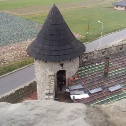 Widok z góry na dach wieży zamkowej w trakcie prac, z widocznymi łatami, częściowo ułożonymi dachówkami i kominem, na tle pól i drogi.