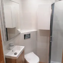 Nowoczesna łazienka z białymi płytkami, drewnopodobną szafką pod umywalką, wiszącą toaletą i kabiną prysznicową z chromowanymi detalami.