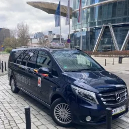 Ciemnoniebieski Mercedes-Benz Vito Taxi z numerem telefonu i herbem na drzwiach, zaparkowany na chodniku przed nowoczesnym budynkiem biurowym w Warszawie.