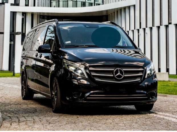 Czarny Mercedes-Benz V-klasa, zaparkowany przed nowoczesnym budynkiem z białymi pionowymi elementami architektonicznymi, widok z przodu lekko z dołu.