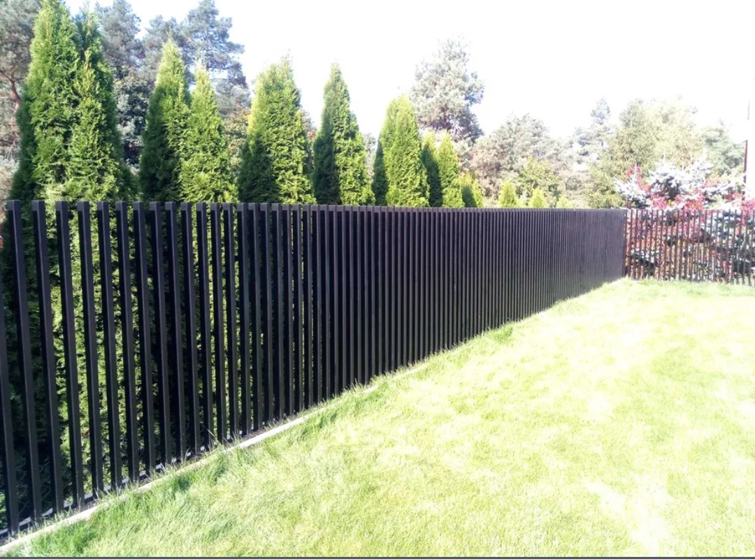 Ciemne, pionowe panele ogrodzeniowe tworzą nowoczesną barierę na tle zielonych tuj i trawnika, z widocznym fragmentem drugiego ogrodzenia w tle.