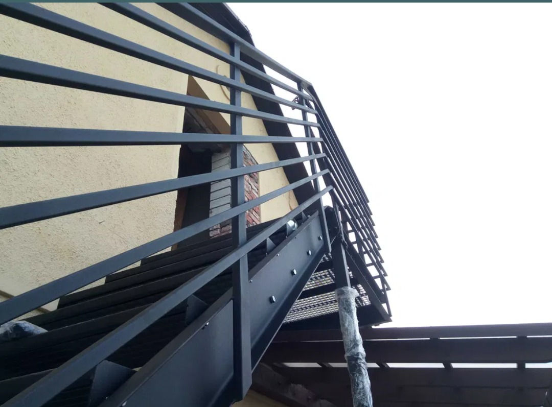 Zewnętrzne schody metalowe z balustradą, przymocowane do elewacji budynku, widok z dołu.