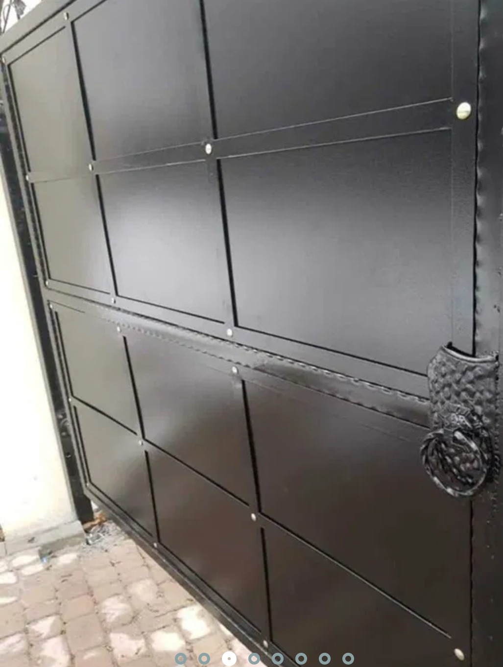 Częściowo otwarta, czarna, metalowa brama wjazdowa z kwadratowymi panelami i ozdobną klamką, widoczna z bliska.