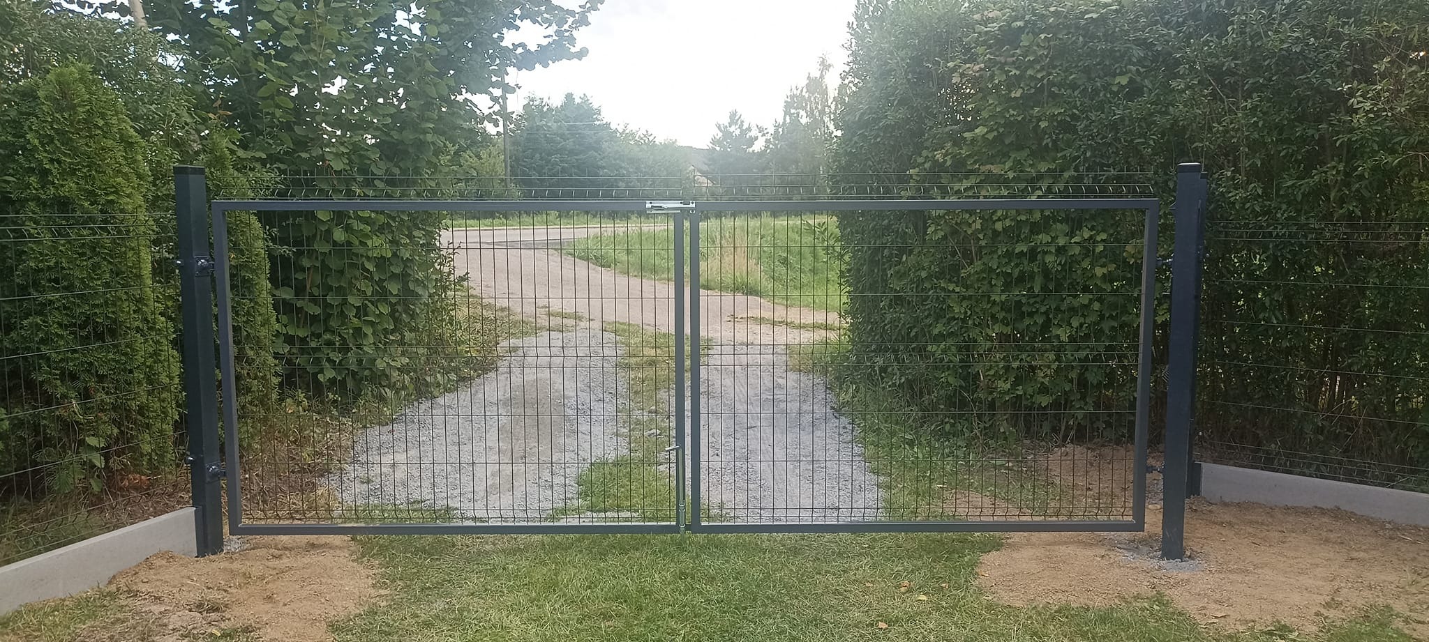 Nowoczesna dwuskrzydłowa brama z metalowej siatki na posesji z widokiem na żwirową drogę wjazdową, otoczona zielenią.