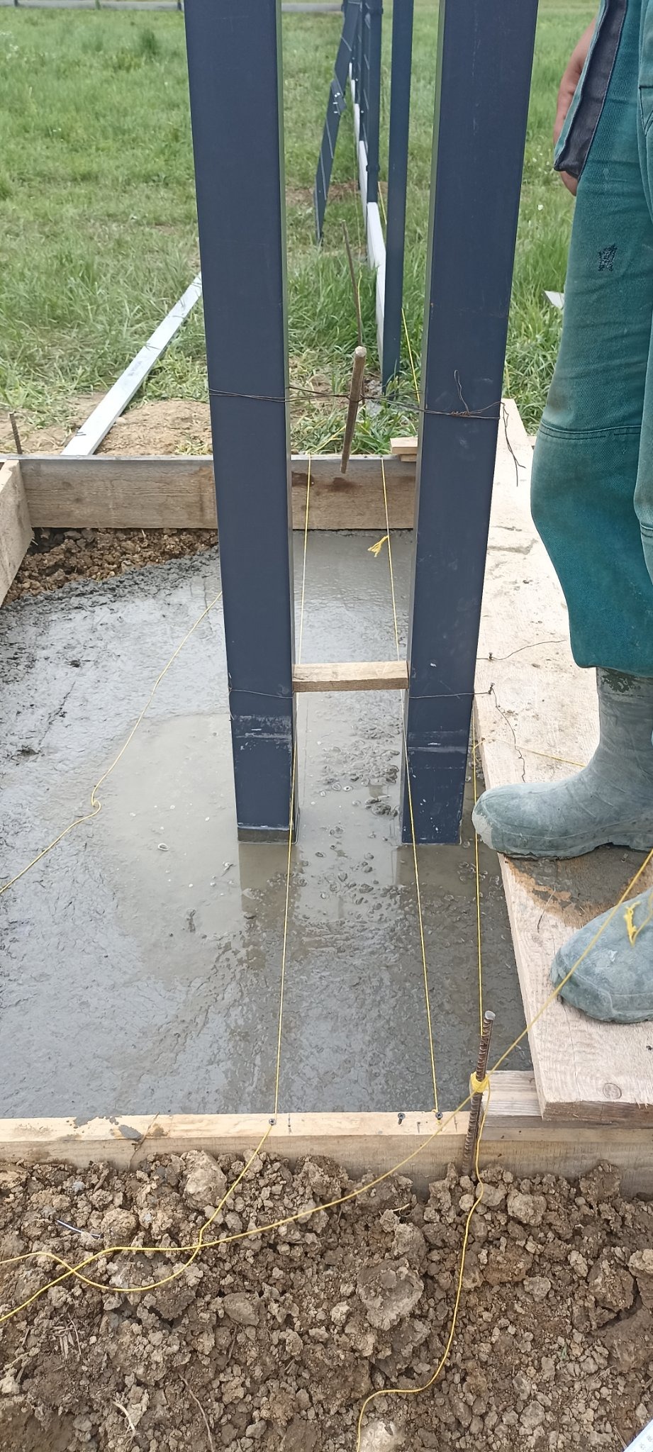 Betonowanie słupków pod konstrukcję, widoczne sznurki wyznaczające linie, drewniany szalunek i fragment roboczej odzieży