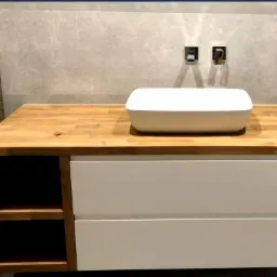 Łazienka z blatem z jasnego drewna i umywalką nablatową, białe fronty szuflad, otwarte półki po lewej stronie, szara ściana z bateriami.