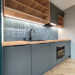 Nowoczesna kuchnia z szarymi szafkami, drewnianym blatem i niebieskimi płytkami w pionowe paski, oświetlona listwą LED pod wiszącymi szafkami z otwartymi półkami.