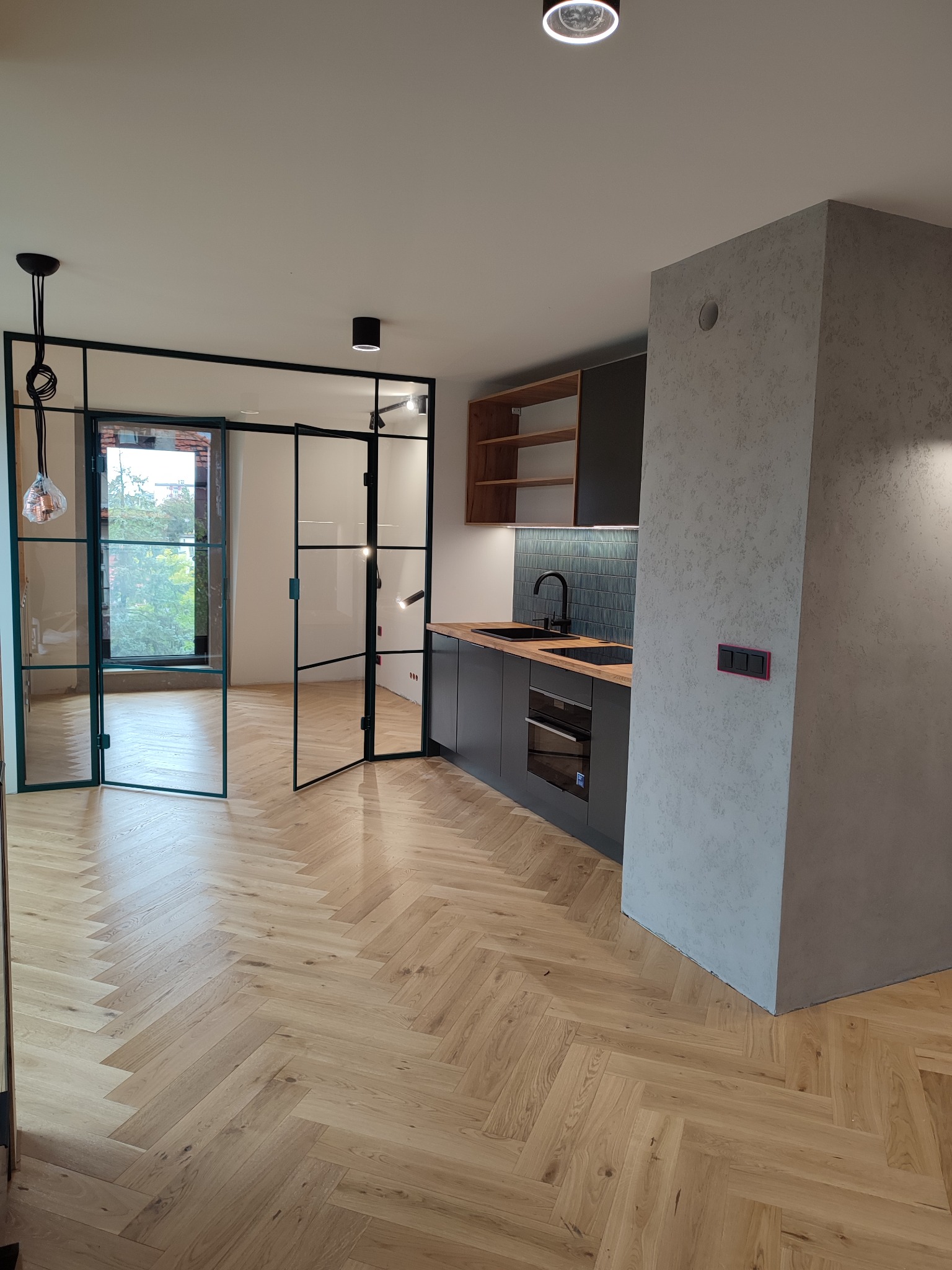 Nowoczesna kuchnia z ciemnymi szafkami, drewnianym blatem i podłogą w jodełkę; przeszklone drzwi z czarnymi ramami oddzielają przestrzeń, a dekoracyjny tynk na ścianie dodaje tekstury.