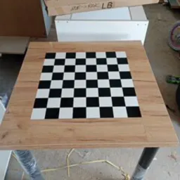 Kwadratowy stolik z blatem imitującym szachownicę, otoczony ramą z płyty drewnopodobnej. W tle kartonowe pudło i biała szafka, całość w warsztacie lub magazynie.