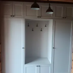 Jasnoszara zabudowa meblowa z szafkami górnymi, wnęką z haczykami i dolnymi szafkami, w drewnianym wnętrzu z parkietem w jodełkę.