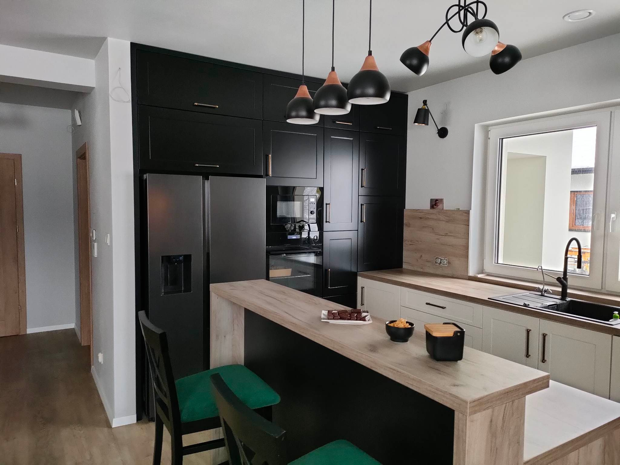 Nowoczesna kuchnia z czarnymi szafkami, drewnianym blatem barowym i zielonymi krzesłami, oświetlona wiszącymi lampami w stylu industrialnym.