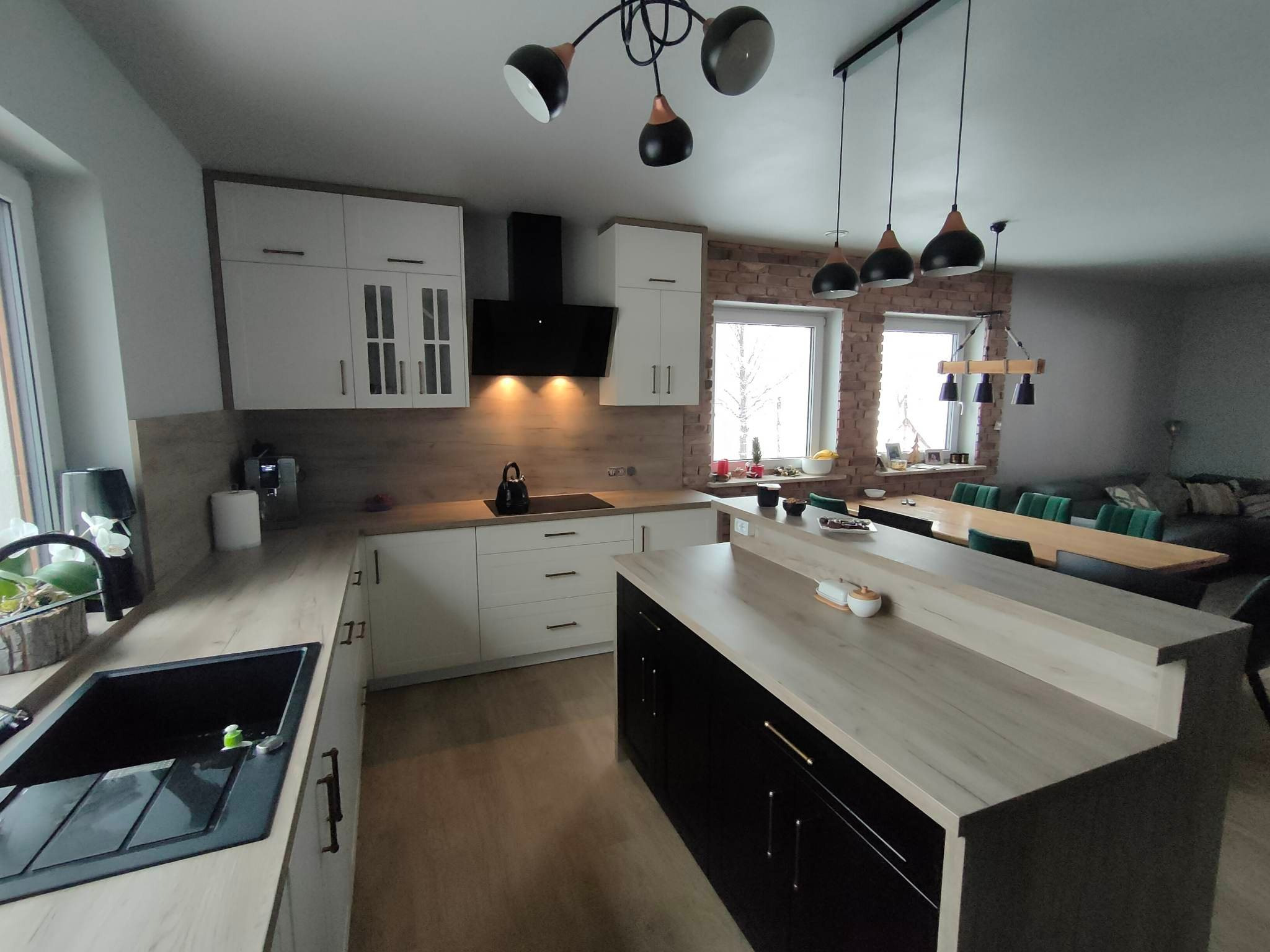 Nowoczesna kuchnia z wyspą, białe szafki z drewnianymi akcentami, czarny okap i wyspa z czarnymi frontami, widok na jadalnię i salon.