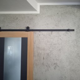 Jarosław Nowakowski - Szklane drzwi przesuwne w drewnianej ramie z czarnymi elementami, zawieszone na czarnej szynie z rolkami, na tle ściany o szarej, chropowatej fakturze.