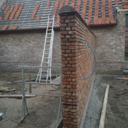 Ceglany mur oporowy w trakcie budowy, widoczne przymocowane rury instalacyjne, w tle rusztowanie i częściowo ułożone dachówki na budynku.