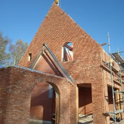 Wznoszenie ścian domu z czerwonej cegły, widoczne otwory okienne i drzwiowe, rusztowanie przy ścianie, żuraw budowlany w tle na tle błękitnego nieba.