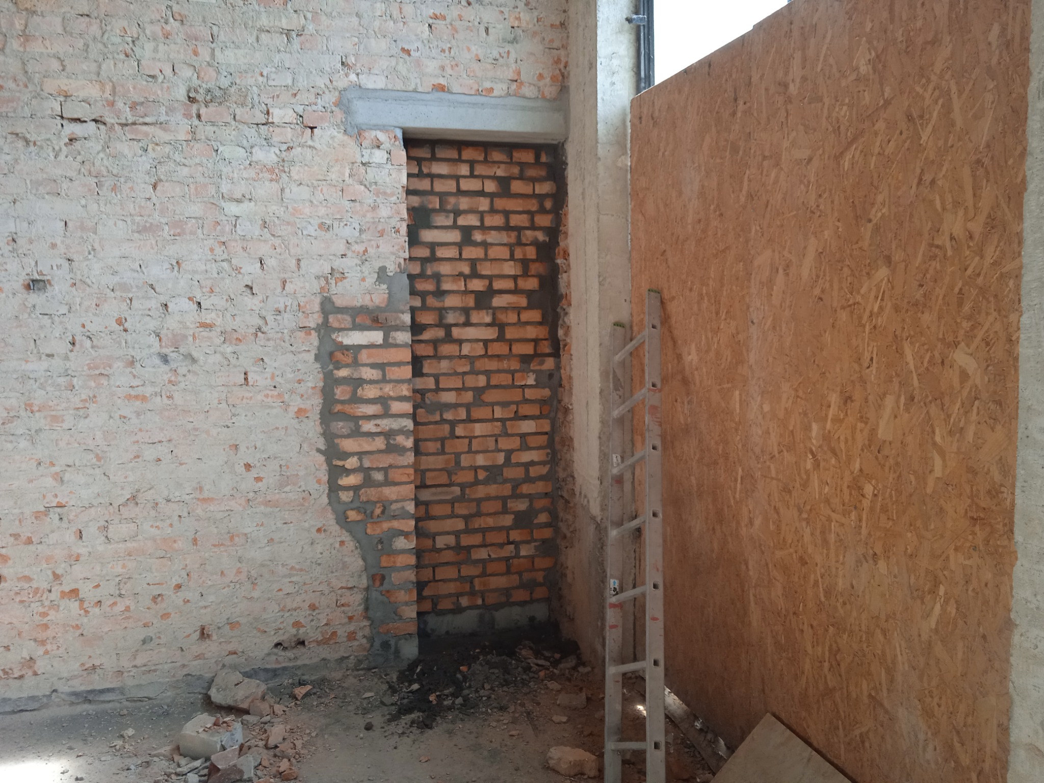 Wnętrze w trakcie prac budowlanych: częściowo otynkowana ściana z cegły, zamurowany otwór, płyta OSB i aluminiowa drabina oparta o ścianę, gruz na podłodze.