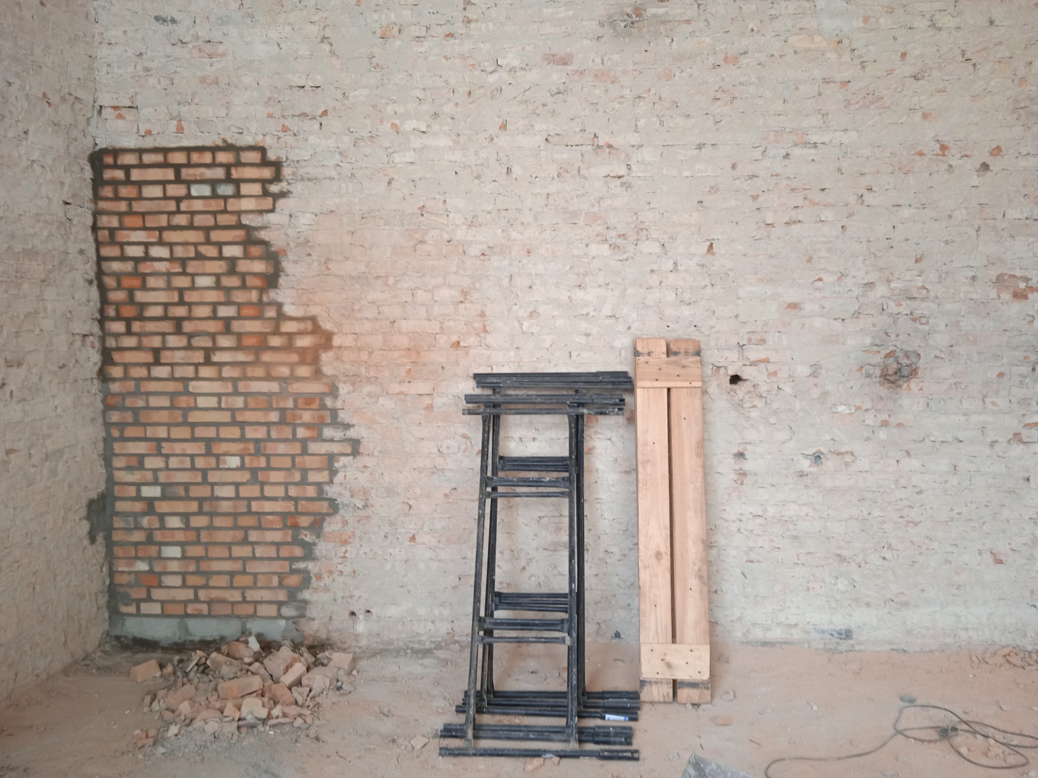 Fragment ściany z czerwonej cegły, częściowo otynkowanej, z widocznymi pozostałościami gruzu, metalowe rusztowanie składane i drewniane deski opierają się o ścianę.