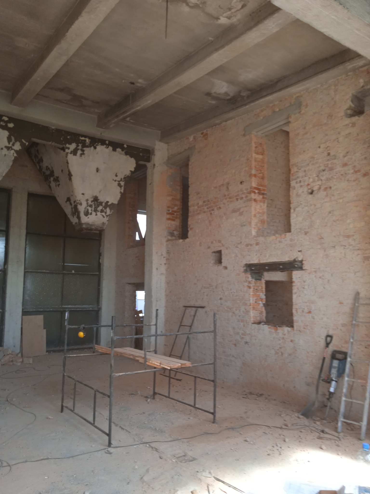 Wnętrze budynku w trakcie przebudowy: surowe ceglane ściany z otworami okiennymi, betonowy strop z belkami, metalowe rusztowanie z drewnianymi deskami, drabina oparta o ścianę, oraz młot udarowy...