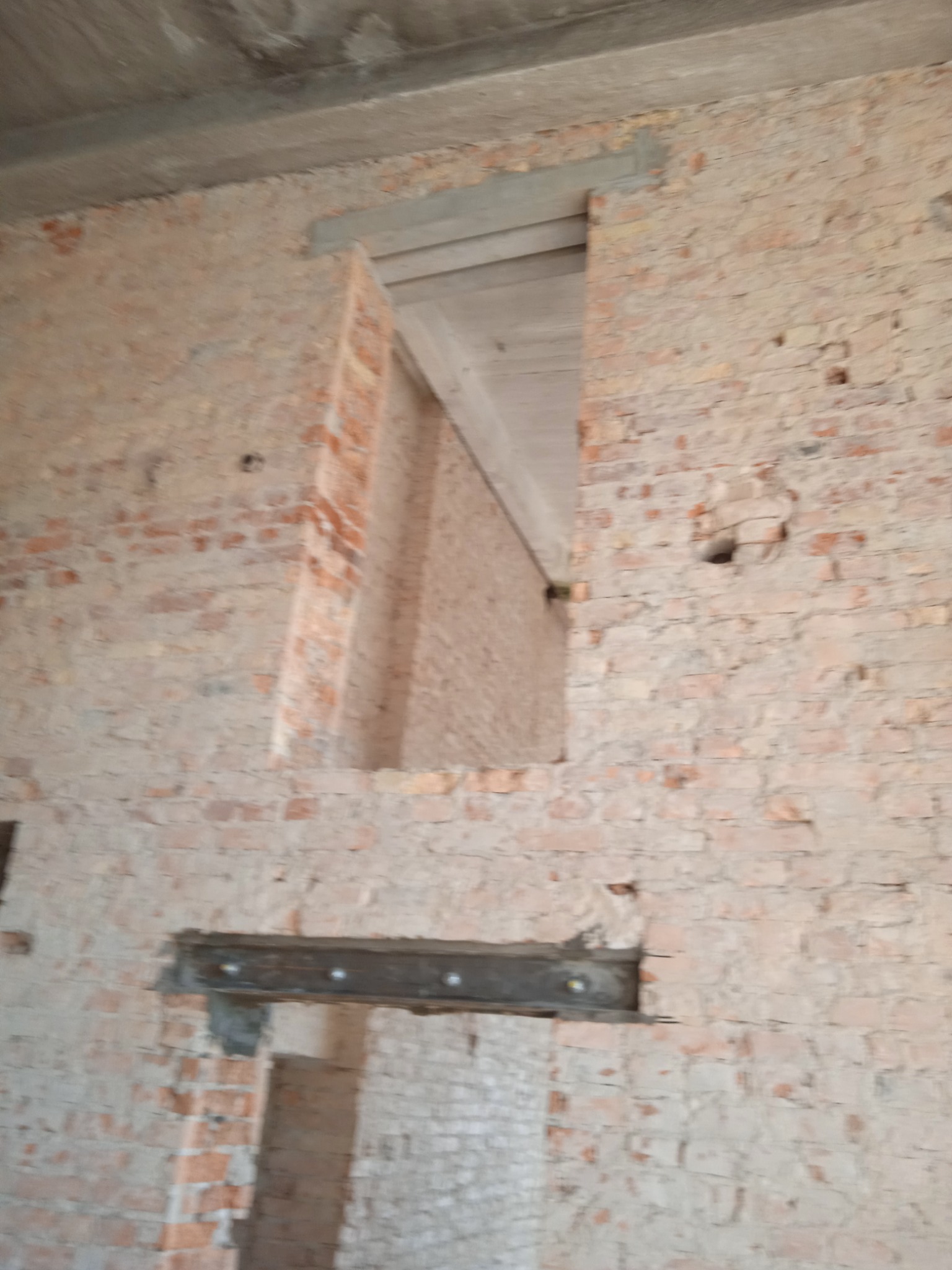 Wnętrze budynku w trakcie prac murarskich, widoczna ściana z cegły z otworem okiennym i stalowym nadprożem, surowy betonowy strop w tle.