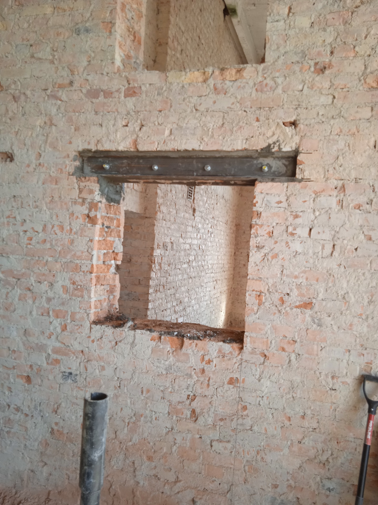Wzmocnienie otworu w ceglanej ścianie stalową belką z widocznymi śrubami, fragment rozbiórki z widocznym fragmentem szpadla i rury.