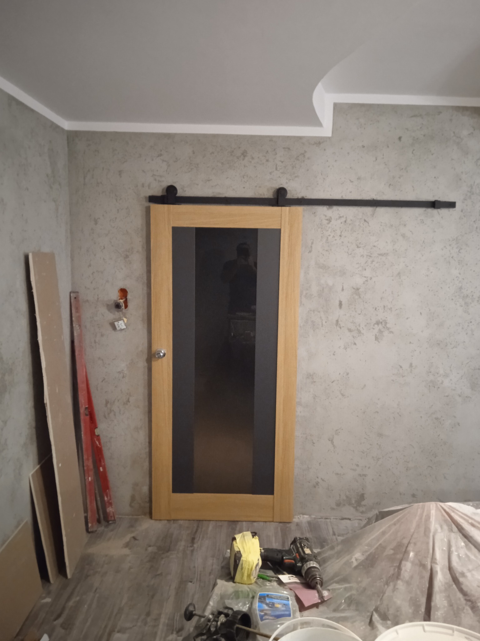 Montaż przesuwnych drzwi wewnętrznych z drewnianą ramą i czarnym szkłem na szynie, widoczne narzędzia i materiały budowlane w nieukończonym pomieszczeniu z teksturowaną ścianą.