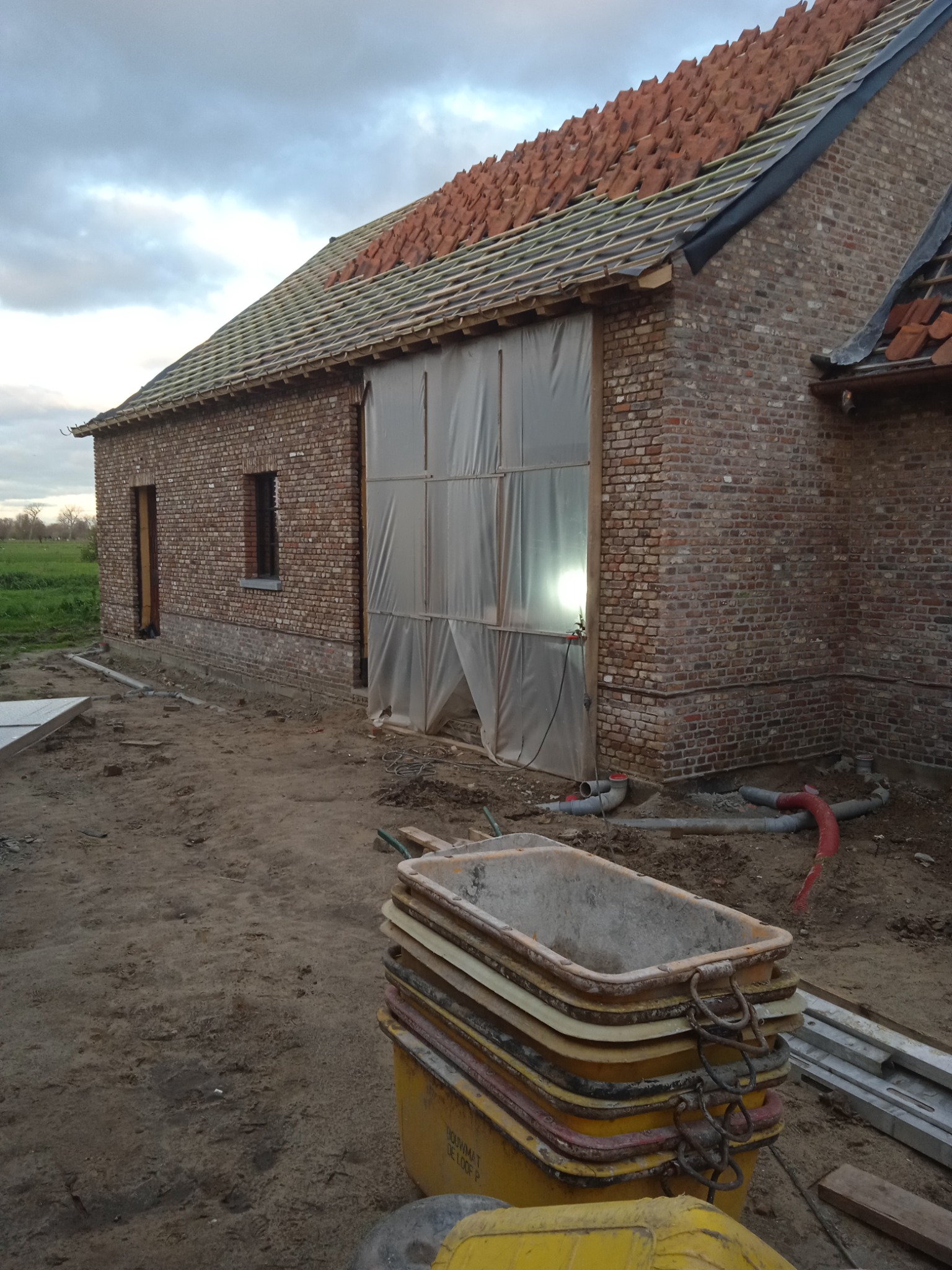 Budowa domu z cegły w Poznaniu w trakcie realizacji, widoczne murowane ściany, częściowo ułożona dachówka, folia budowlana osłaniająca otwór oraz sterta żółtych pojemników budowlanych.
