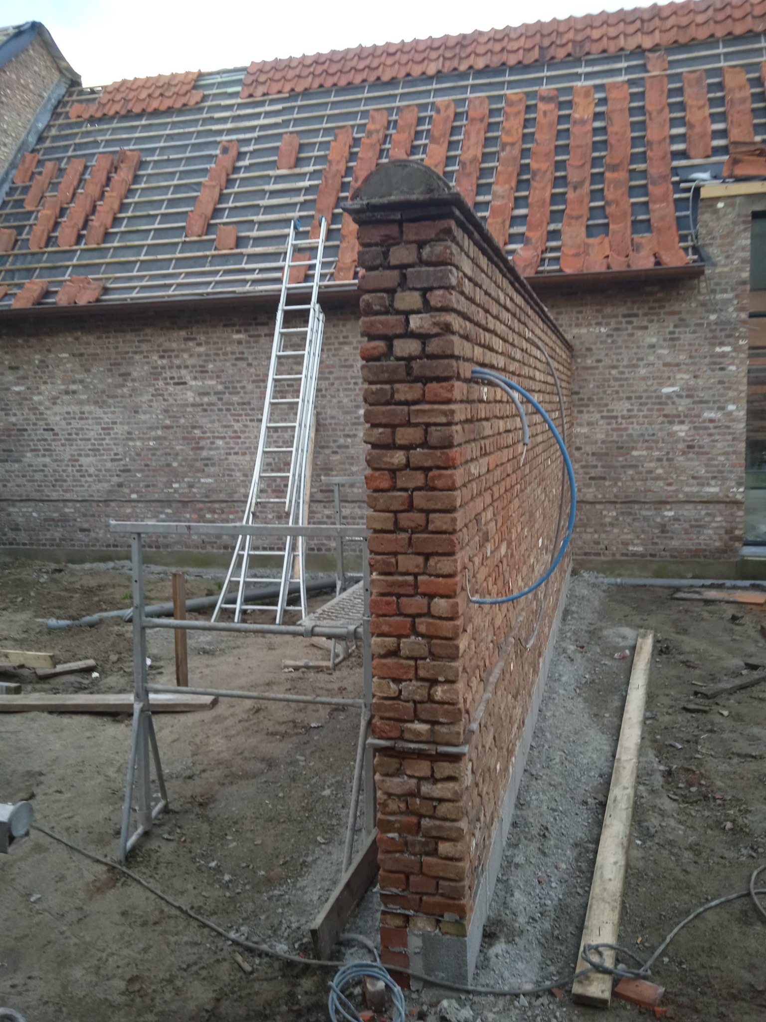 Ceglany mur oporowy w trakcie budowy, widoczne przymocowane rury instalacyjne, w tle rusztowanie i częściowo ułożone dachówki na budynku.
