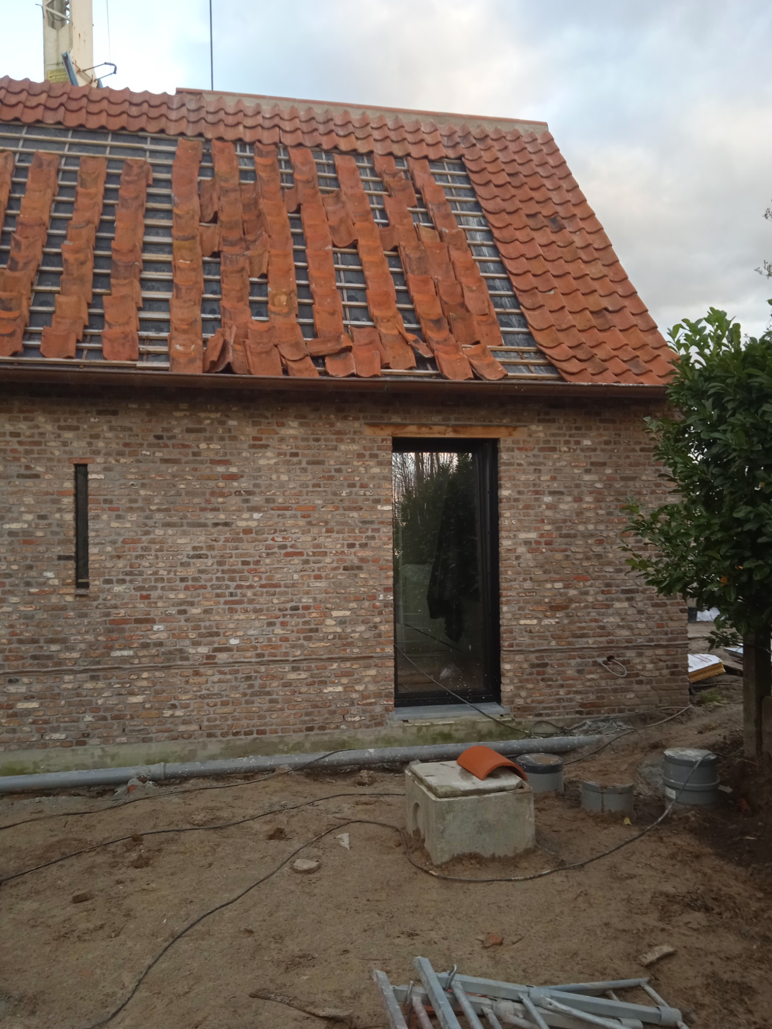 Budynek w trakcie remontu dachu z częściowo ułożoną czerwoną dachówką, widoczna murowana ściana z oknem i elementami instalacji zewnętrznej.