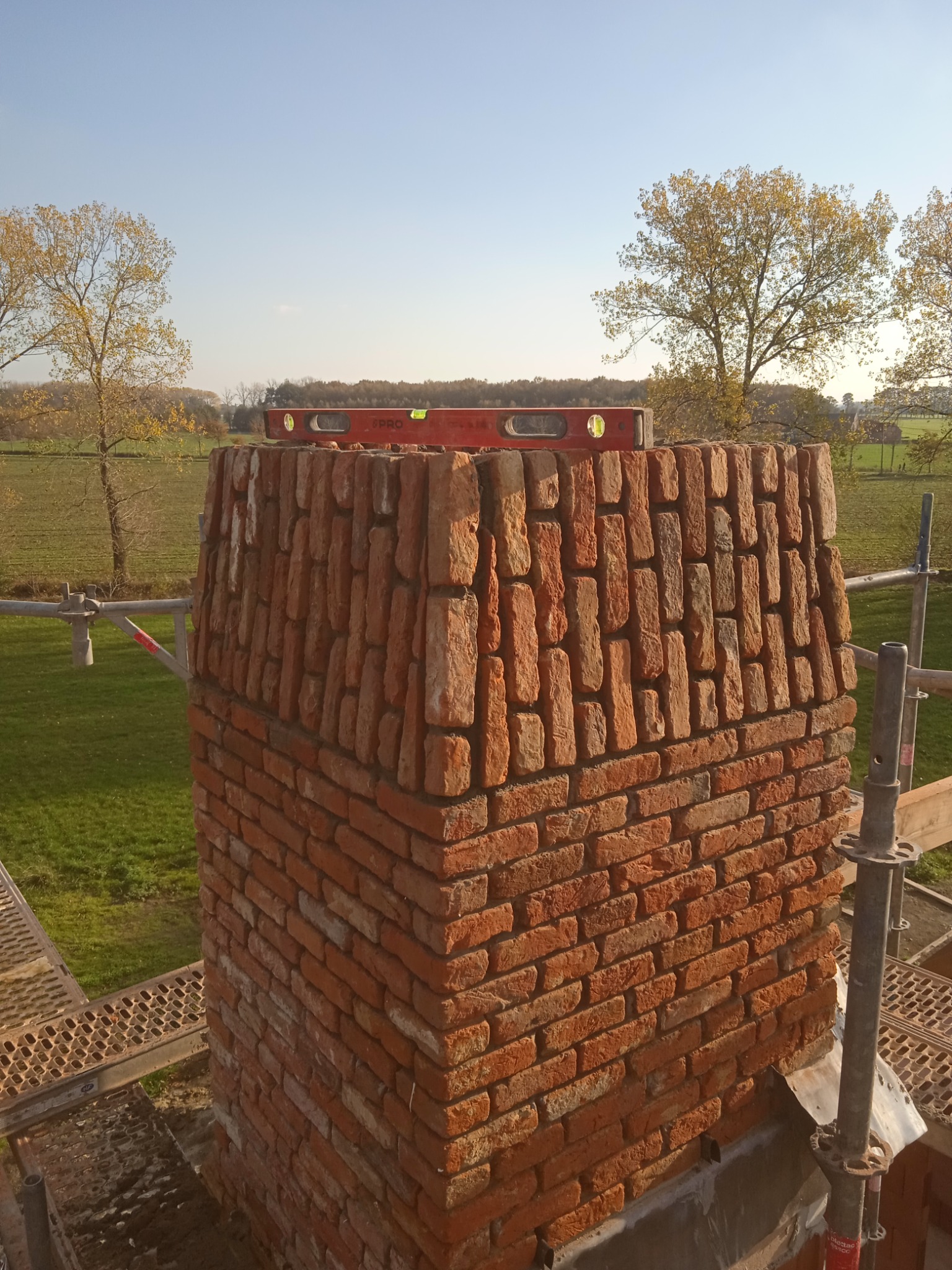 Wznoszenie komina z czerwonej cegły na tle zielonego pola i drzew, z użyciem poziomicy dla zachowania pionu.