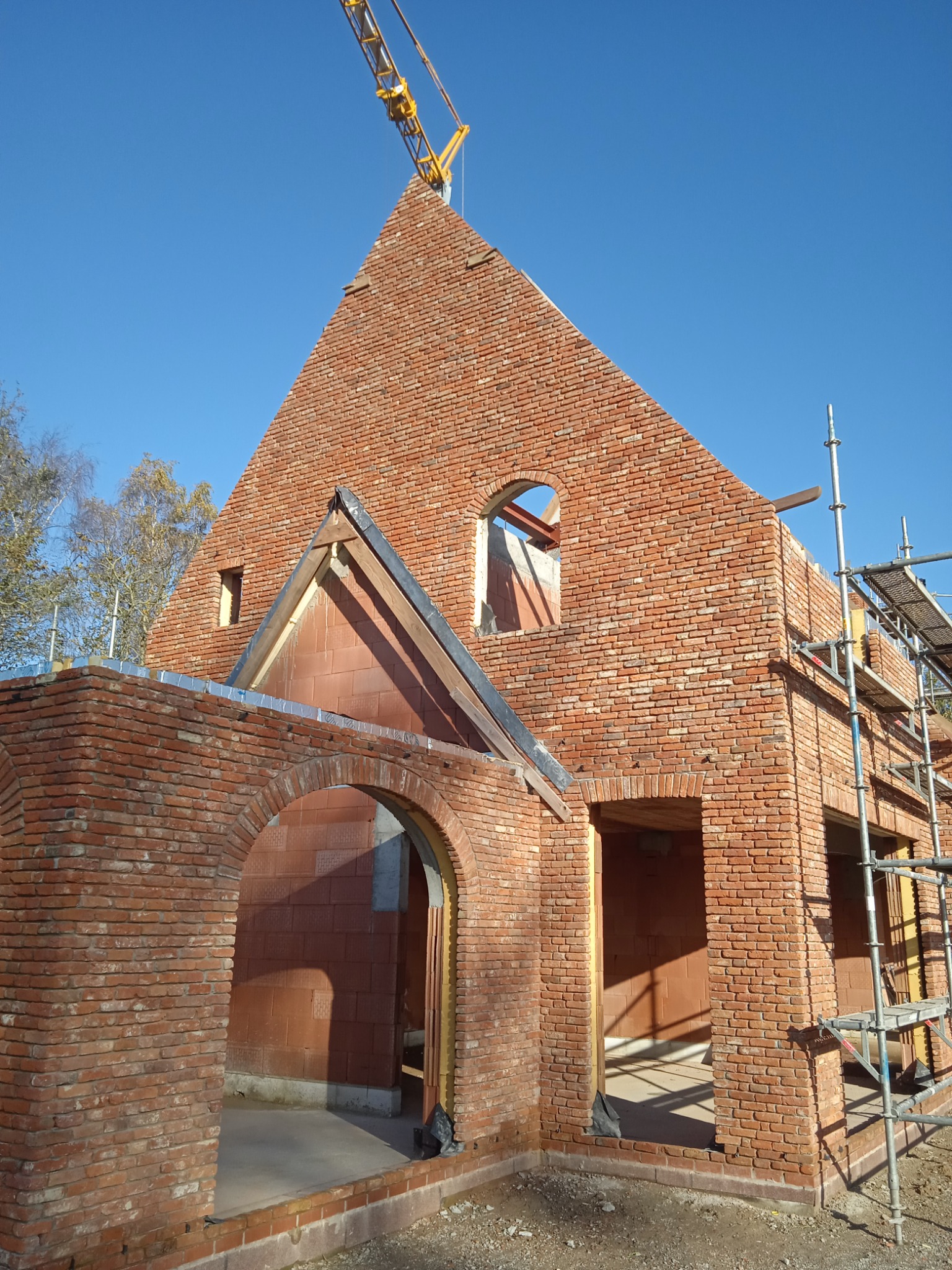 Wznoszenie ścian domu z czerwonej cegły, widoczne otwory okienne i drzwiowe, rusztowanie przy ścianie, żuraw budowlany w tle na tle błękitnego nieba.