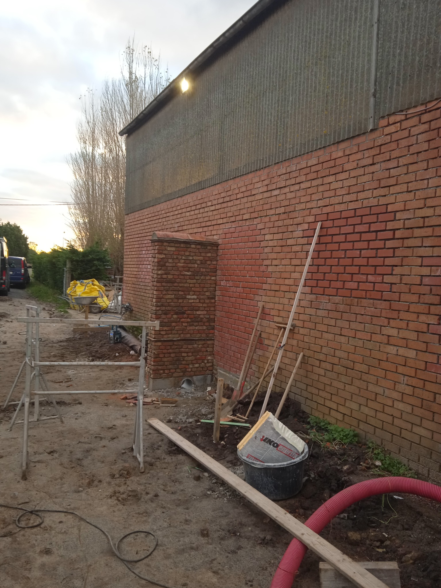 Murowany fragment ściany budynku z czerwonej cegły, częściowo pokrytego blachą falistą, z widocznymi narzędziami murarskimi, rusztowaniem i materiałami budowlanymi na ziemi.