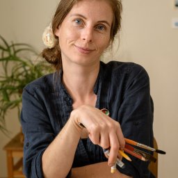 ENEN STUDIO - Portret uśmiechniętej kobiety z pędzlami w dłoni, siedzącej przy stole z dwoma szkicami akwarelowymi.