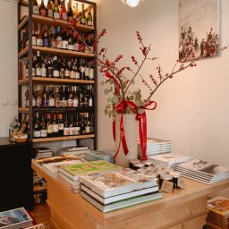 ENEN STUDIO - Wnętrze sklepu z winami i książkami, regał z butelkami wina w tle, na pierwszym planie drewniany stół z wazonem z czerwonymi gałązkami i stosem książek o tematyce kulinarnej i ogrodniczej, małe...