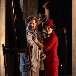 ENEN STUDIO - Kobieta w czerwonym kostiumie poprawia klapę marynarki mężczyzny stojącego przed sztalugami w stylizowanej scenerii teatralnej z rzeźbami w tle.